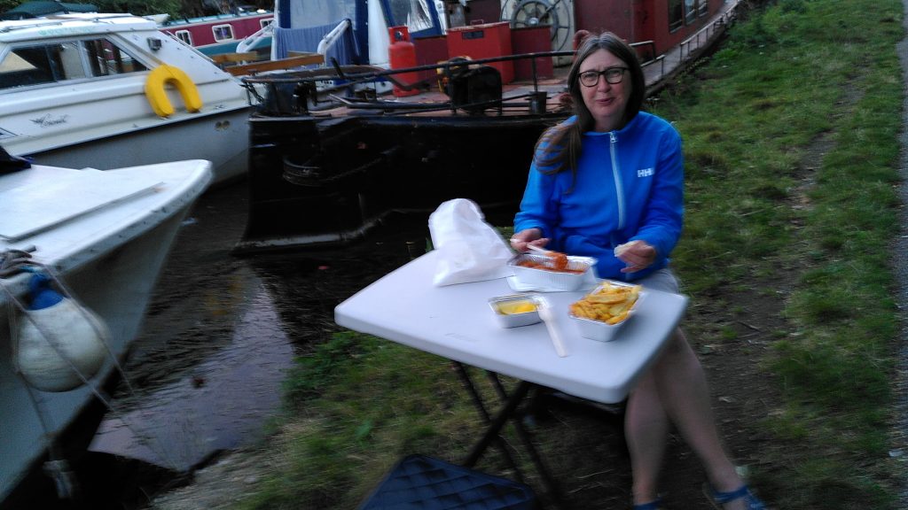Clare at a towpath table.  The small table has a stool either side and portions of food in foil trays and paper bags.  The canal has two boats moored side-by-side.