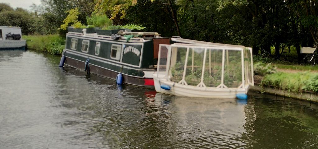 Narrowboat with greenhouse.  Moored as a tender to a narrowboat is a converted rowing boat.  Glass sides have been added to create a floating greenhouse.