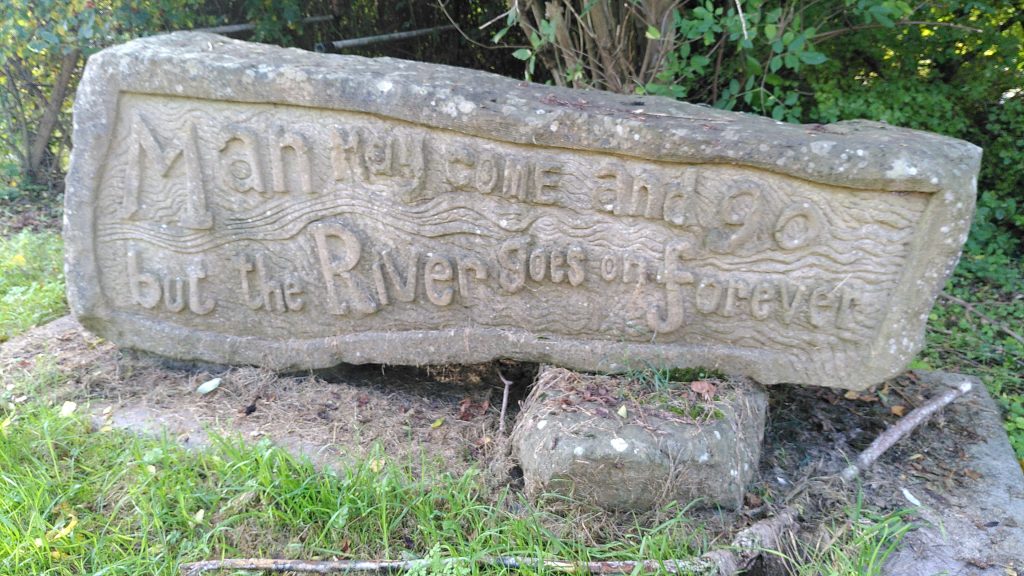 Stone tablet at the side of the river.  The block is carved in an uneven style with the words "Man may come and go but the River goes on forever".