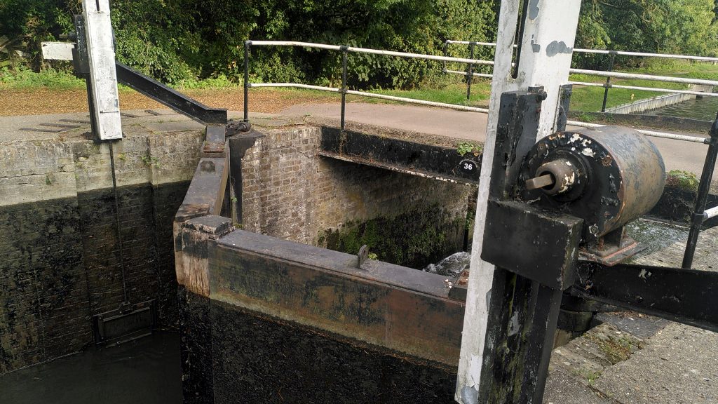 Tail gates of a River Stort Lock.  In the foreground, the hydraulic winding gear spindle can be seen.  Across the canal the raised paddle can be seen underneath the corresponding gear. On the other side of the lock gate water rushes out of a tunnel.