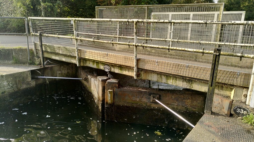Tail gates of a River Stort Lock. A road bridge immediately downstream has had a footbridge built alongside it. The bridge crosses above the gate hinges where the arms might normally be attached. A hydraulic mechanism has been installed to allow the gates to be operated.