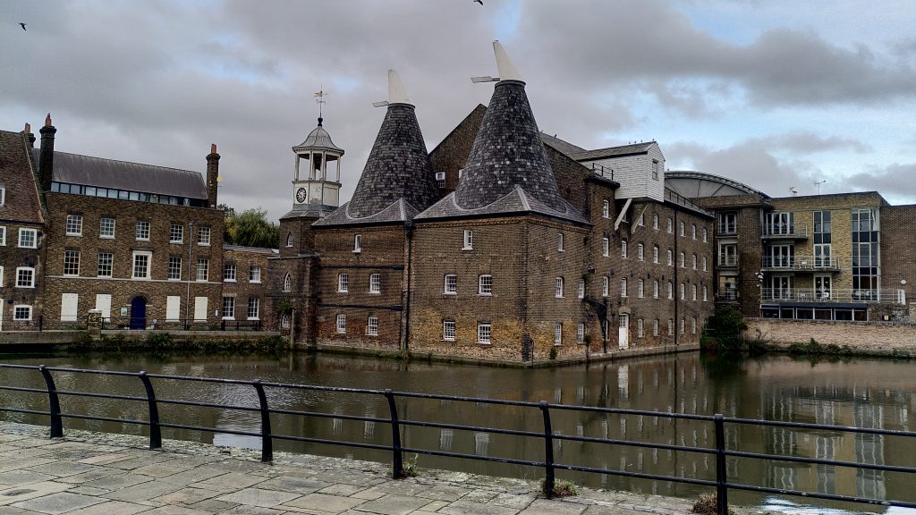 Mill Building at Three Mills.  A four storey brick building with rows of white painted windows.  The slate roof forms two pyramids with white cones at their peaks and a clock tower alongside. A railing at separates the path we're standing on from the tidal river below.