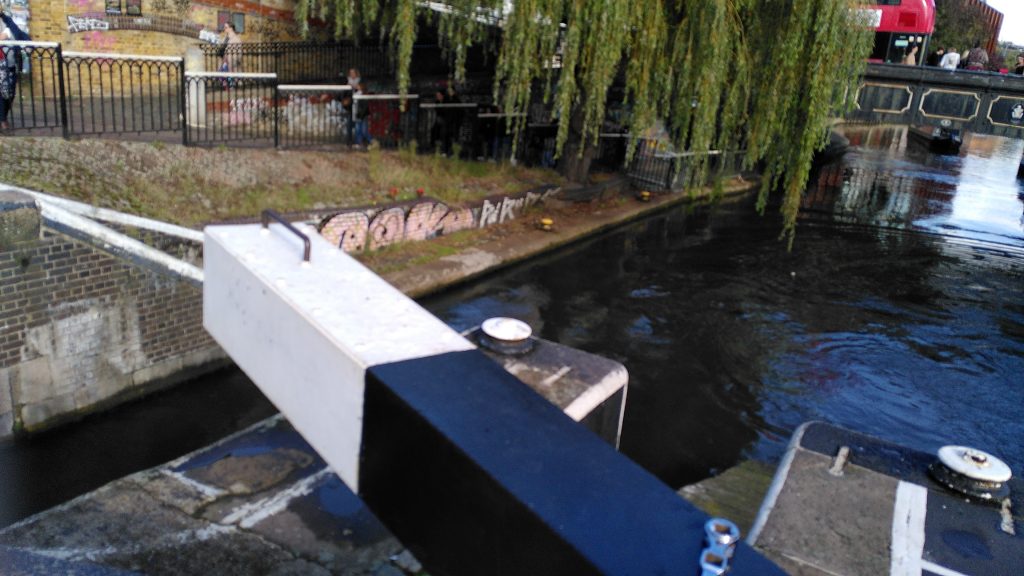 Lock arm viewed from the hinge. The arm is painted white for the last few feet and black elsewhere.  White paint also marks the edge of a set of steps sunk in to the wall beneath the arm.  The steps are not visible.  There is a platform at the edge of the canal six feet below.   A narrowboat is just visible under a bridge further down the canal.