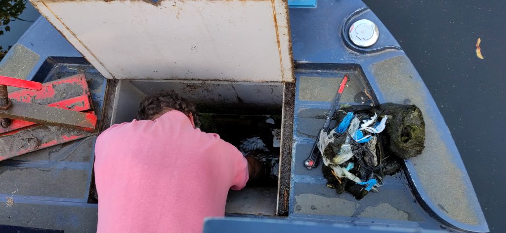 The back and shoulders of someone with their head in the weed hatch.  The cover of the weed hatch is open and a collection of detritus has been dumped on the rear deck.