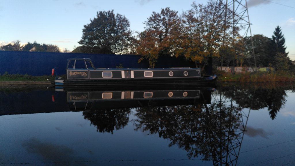 A narrowboat moored at the side of a canal.  There are trees on the bank behind the boat and the sky above is blue.  The scene is reflected in the still waters of the canal.