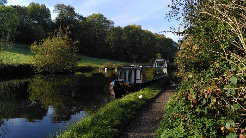 Narrowboat moored on the canal side.  The towpath has a hedge alongside it and a grass verge next to the canal.  The far side of the canal is a gently sloping field rising to woodland.  The sky above is blue.