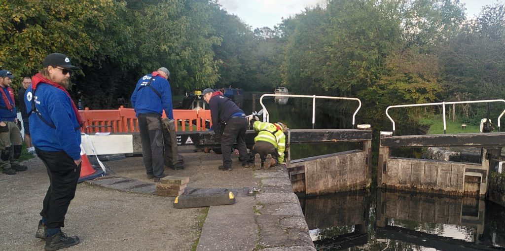 Workers preparing to reseat a lock gate.  The near side lock gate is at the wrong angle.  The arm is barriered off while workers place a jack underneath preparing to reseat it.