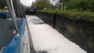 View down the side of a narrowboat in a lock. Most of the surface of the lock is covered in a white foam several inches deep.