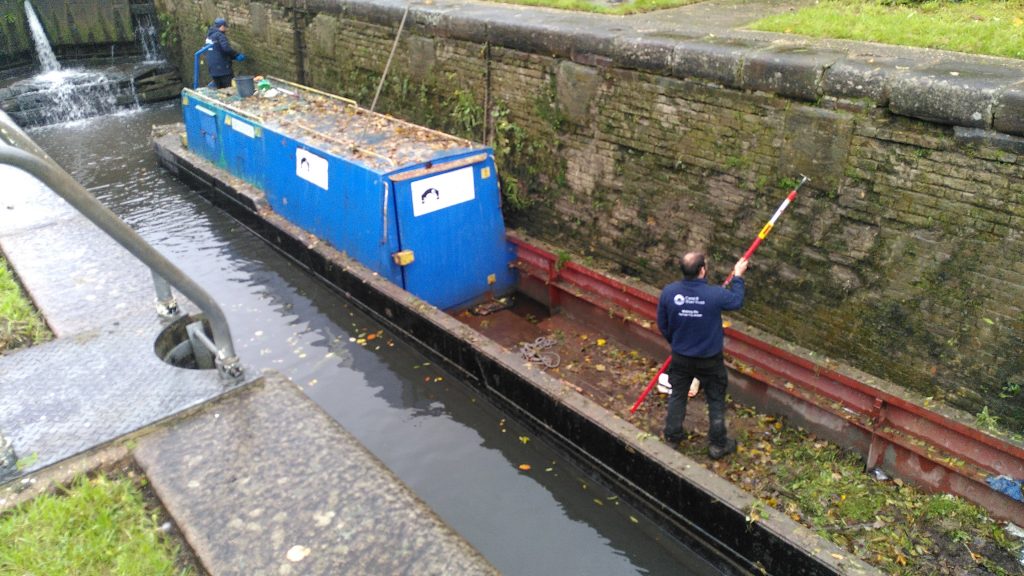 Work boat in a lock.  Two workers are removing vegetation from the walls inside a lock using the boat as a platform.  The boat is a narrowboat with a small cabin at the back and an open deck at the front.