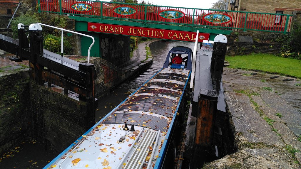 A narrowboat entering a lock under a bridge.  Only one gate is open but the narrowboat fits neatly.  A low bridge just clears the cover over the driver of the narrowboat.  The bridge is painted in high contrast colours with the words "Grand Junction Canal".  The grey roof of the boat is speckled with autumn leaves in bright yellows and oranges.
