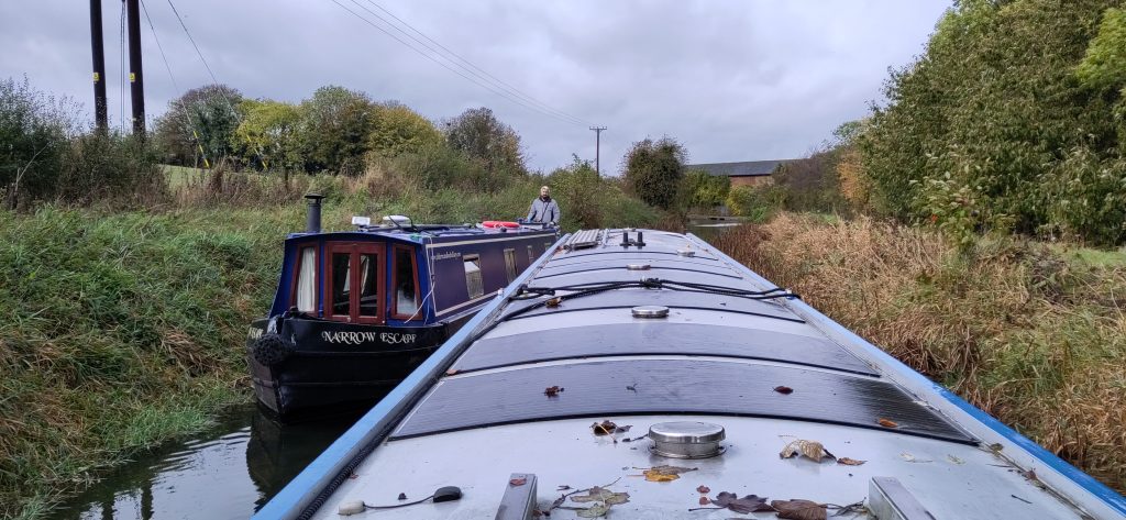 Two narrowboats meet.   Viewed from the stern of a narrowboat, there is no visible canal except in front of the oncoming boat.  The banks on either side are gently sloping vegetation leading to fields.