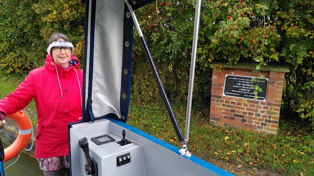 A woman at the helm of a narrowboat. She is wearing a coat and headband to keep warm. Behind her on the towpath is a commemorative plaque on a brick pillar. The plaque records that the arm was opened in 2004 by the legacy of Tim Wilkinson.