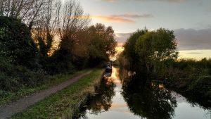 Sunset over a canal. The view is straight down the canal with a towpath running along one side. There are trees and bushes beyond the towpath and on the opposite bank. Through the trees there are clouds glowing in yellow and orange in a grey-blue sky.