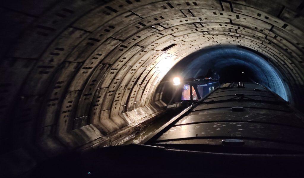 Two narrowboats passing in a tunnel.  The view is from the steering position of one of the boats.  The light at the end of the tunnel can be seen over the roof straight ahead.  The tunnel walls are made of jointed rings of concrete illuminated by the light of the oncoming boat.