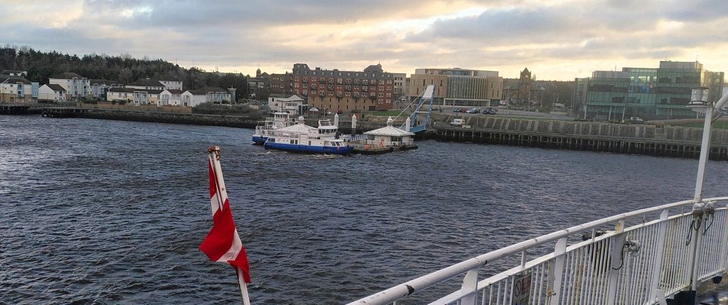 Ferry from South Shields.  A small ferry is docked at a jetty below a line of buildings at the side of a river.  We are looking from the deck of a ship which is obviously much larger.  The larger ship is flying a Danish flag.