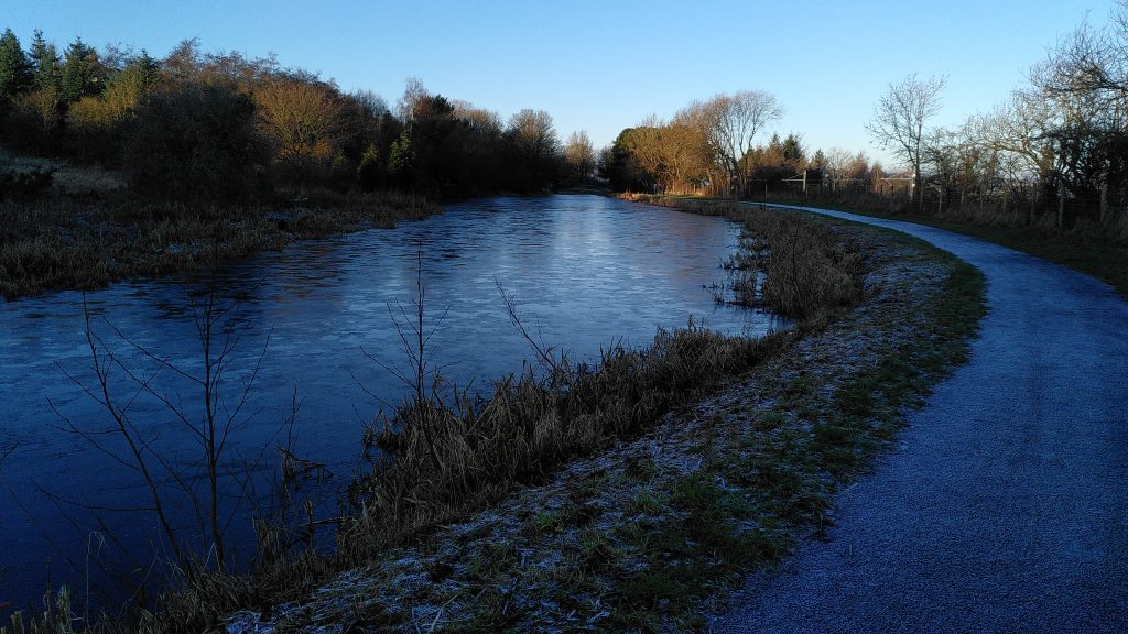 A view over a wide section of the Union Canal.  The towpath is covered in frost, and the water surface is covered in ice.  The sky is blue and cloudless with sun shining on the bushes further along the water.