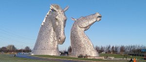 Kelpies. Two sculptures of the head and neck of horses. Each is a hundred feet high. The sun is glinting off the metal , the sky is bright blue behind them.