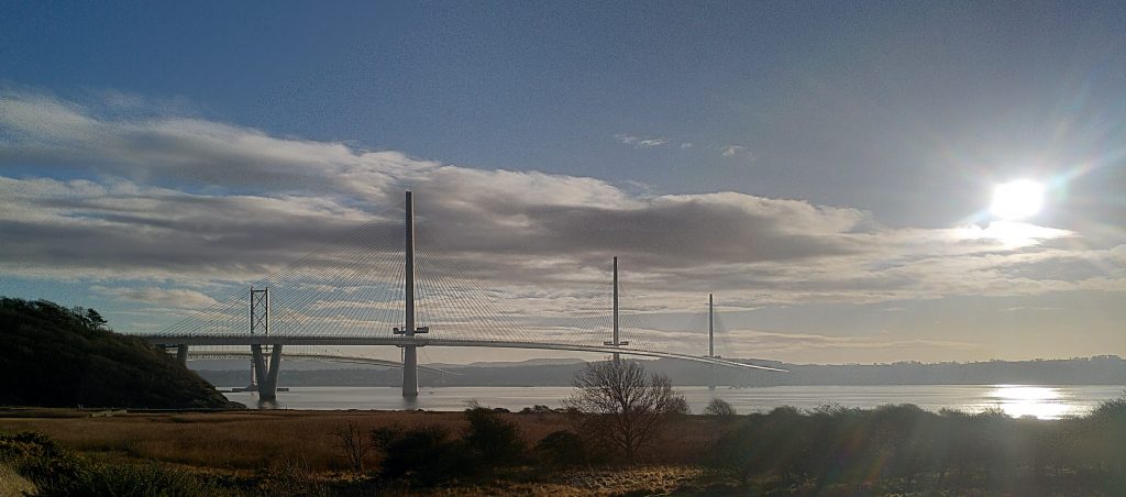 Queensferry Crossing.  The sun is reflected on the waters of the Forth.  The Queensferry Crossing with its three towers largely obscure the older Forth Road Bridge beyond.  The sky is blue but with a bank of low cloud visible.