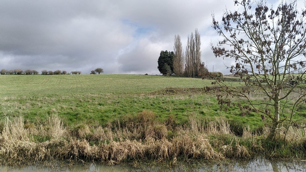 View across a canal to a large gently sloping field of short grass.  There are spindly trees on the horizon with grey clouds in the sky. 