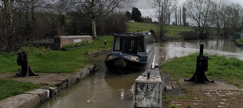 A narrowboat entering a lock. We are looking along  the arm of the open top gate.  Resting on the end of the arm is a windlass. The driver is leaning out to see where she is going.