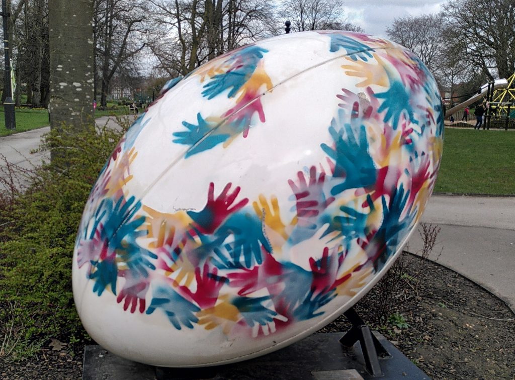 A giant rugby ball sculpture. The sculpture is in a park with grass and trees around. The ball is predominantly white, with stencilled multicoloured handprints forming a band.