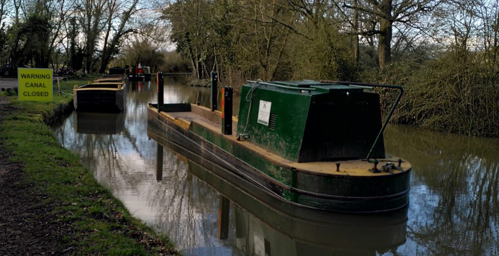 Work boats moored in the canal.  A large yellow sign says "Warning Canal Closed".  The blue sky is reflected in the surface of the canal as it curves away behind some trees.