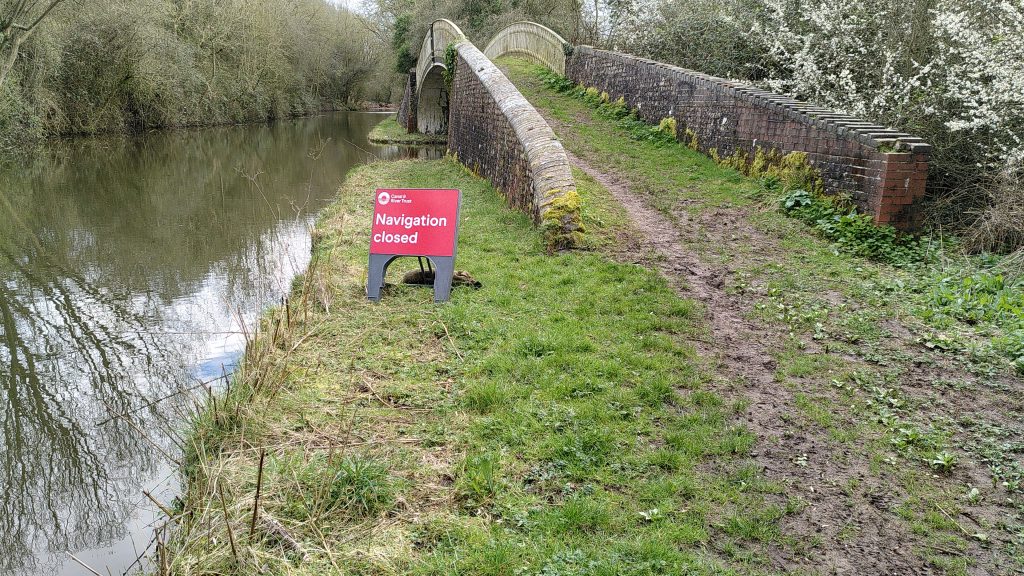 Sign saying "Navigation Closed".  The sign is free standing at the side of the towpath.  Beyond the sign a stone arch bridge takes the towpath over a side branch off the main canal.