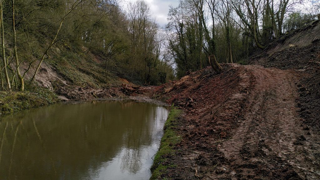 Landslip blocking the canal.  Mud has slithered down from both sides of the embankment forming a blockage several feet above water level.  Stumps of trees stick out from the towpath side.  The towpath is buried to a height taller than a person.