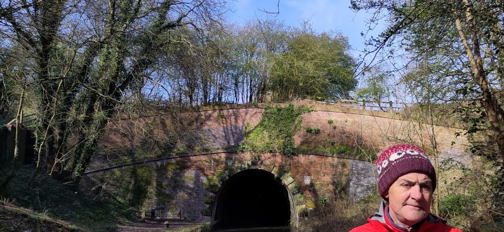 Exiting the tunnel.  The tunnel portal is a dark arch in what looks like a brick bridge with trees growing on top.  The sky above is bright blue and cloudless.  The head of a man in a hat is in the foreground.