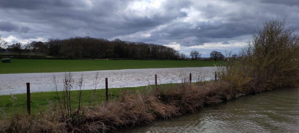 Flooded field.  A field viewed from the back of a narrowboat has a stretch of water in it that seems to be wider than the canal.  There are trees on the far side of the field.  The sky is filled with ominous looking clouds.