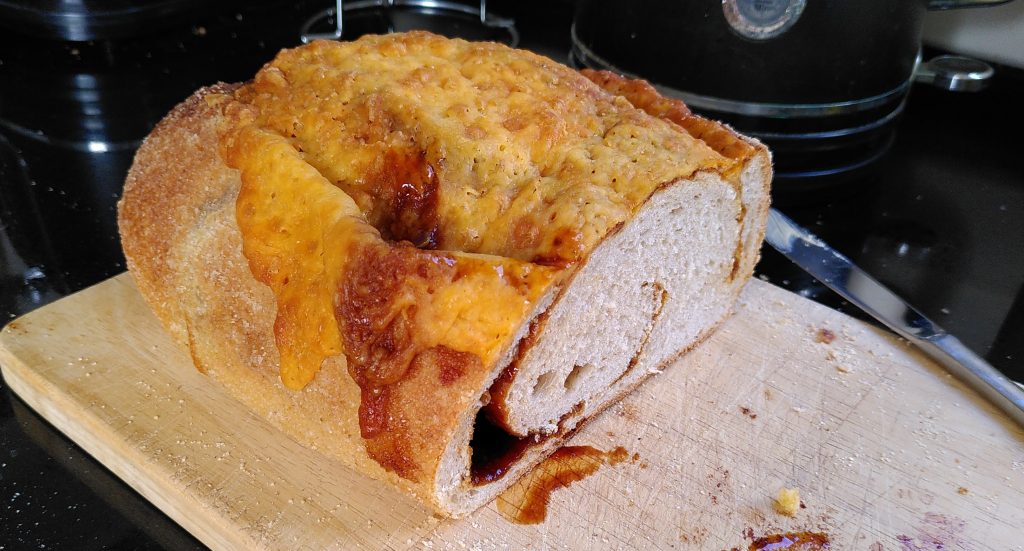 Cheese and marmite loaf.  Half of a loaf of bread is sitting on a wooden breadboard.  The top of the bread is covered in melted cheese.  The cut edge of the loaf reveals that it has a thick layer of marmite in swirls within the bread.