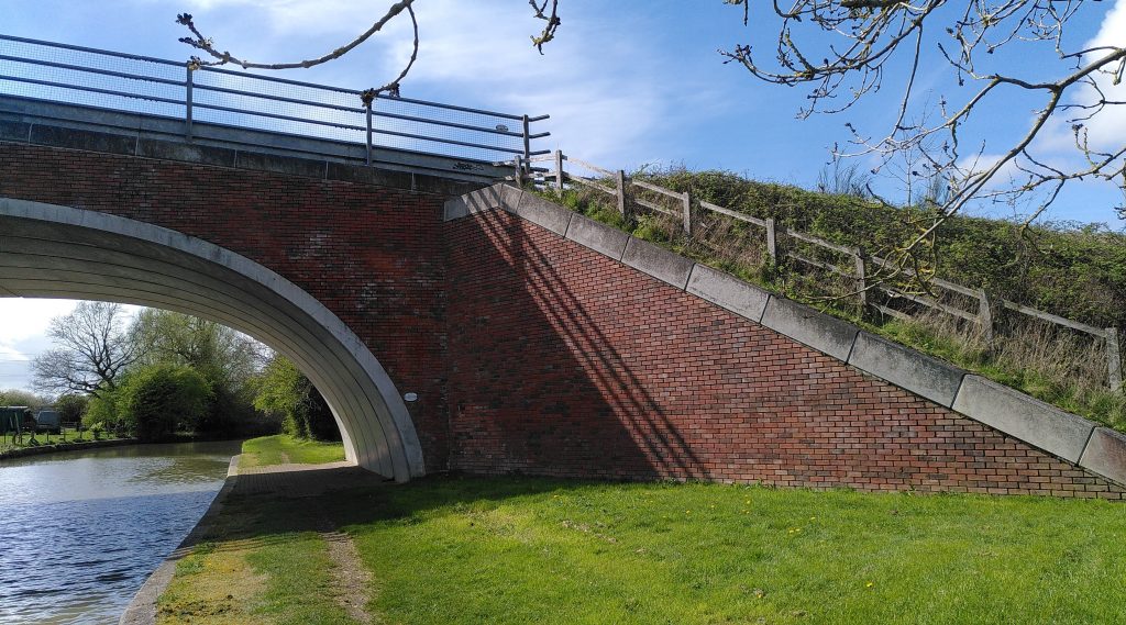 Canal road bridge.  A modern concrete arch bridge with brick facing rises high over the canal.  The retaining wall slopes at a little under 45 degrees towards the towpath.