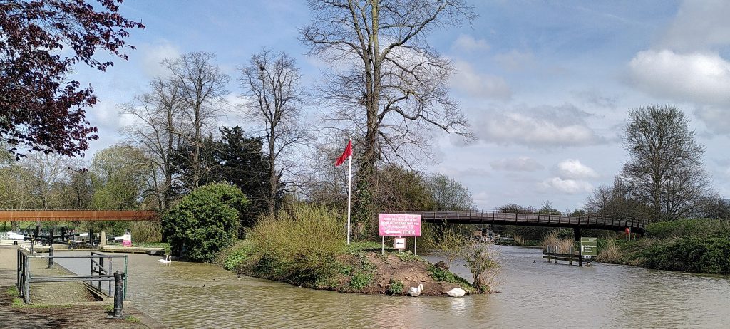 Red flag.  The entrance to the lock channel from the main flow of the river is marked by a large arrow and a red sign.  Next to them is a flagpole with a red flag flying. The lock island is covered in trees and shrubs.  A pedestrian bridge crosses both parts of the river beyond the lock.