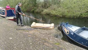 Sofa surfing. A man is using a boat hook to pull a sofa which is floating in the canal. He is in the space between two moored boats having walked around the gunwale of the first and about to go around the second.