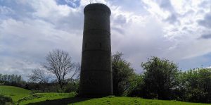 Tunnel chimney. A brick chimney rises out of a small ridge in an empty field. The small trees in the hedgerow behind are dwarfed by the structure. The sky behind is bright with some small dark clouds.