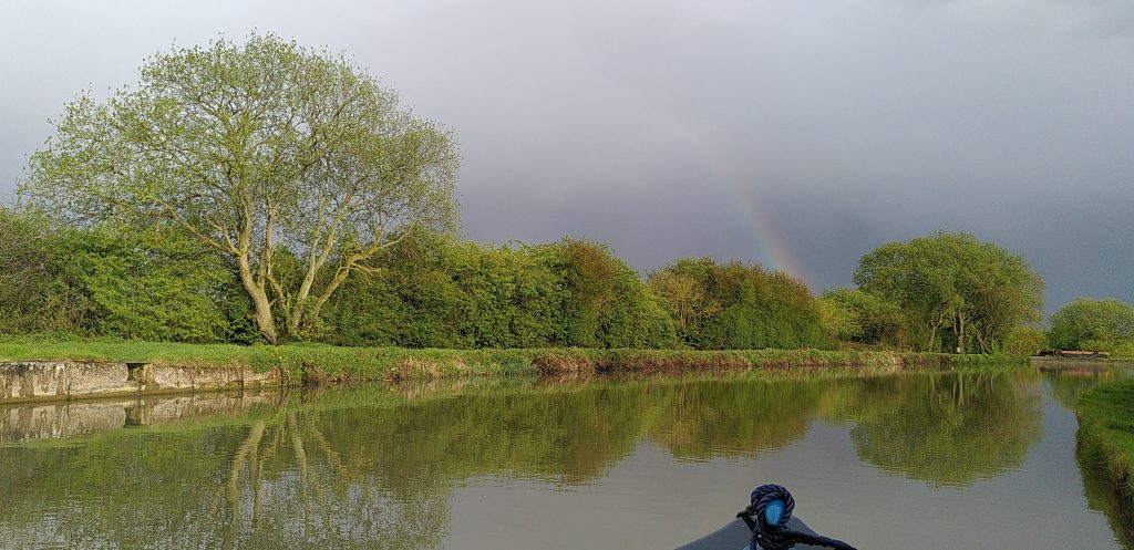 View from the bow. The still water of the canal is bounded by the far bank receding in to the distance. The bank is lined with trees, from which the stub of a rainbow is rising.