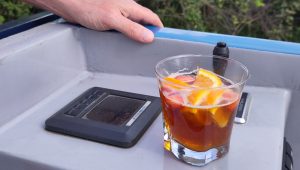 Pimms for the driver. A glass of Pimms with slices of orange and strawberry sits on a shelf. The shelf is at the back of a narrowboat with a control panel and a joystick visible. The drivers hand rests on the edge of the boat, ready to take a sip.