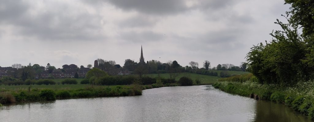 Braunston. A wide section of canal narrows as the canal turns away from the hill ahead. A church spire sits prominently on the ridge. The sky behind is dark and threatening.