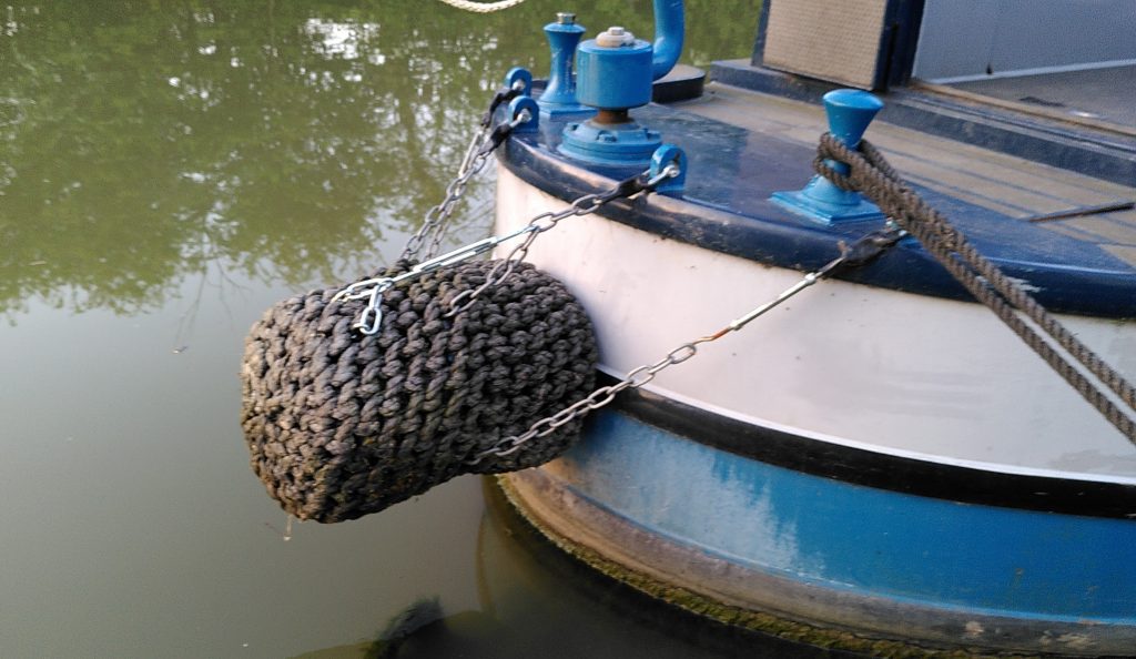 Fender. The stern of a narrowboat with a cylindrical fender in prominence. The fender is held up by a selection of chains. One piece of chain is particularly shiny and is fitted much further back than the rest. Some discolouration of the fender hints that it has previously sat much lower.