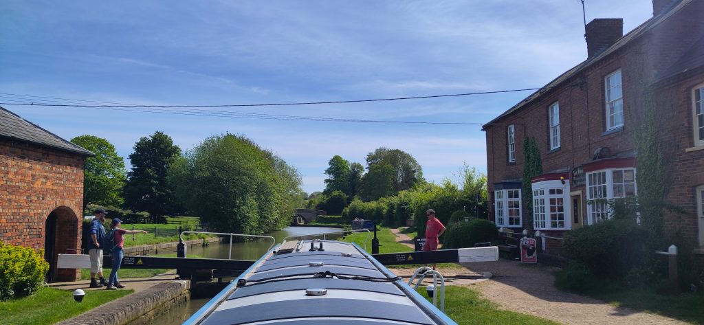 Braunston Bottom Lock.  View from the stern of a narrowboat rising in Braunston Bottom Lock.  There are people on either side of the lock at the head gates.  There are brick building on both sides of the lock.  Ahead the lock has trees and hedges on both sides. 