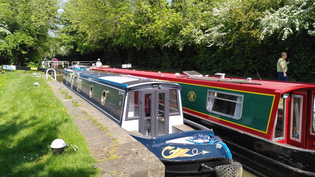Narrowboats in a lock.  Two narrowboats side-by-side in a canal lock.  The gunwales are almost level with the grass at the side of the lock.  Crew at the stern of both boats are in full sunshine, but the canal behind is in the shade of overhanging trees.