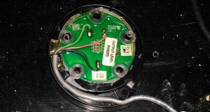 Tank sensor. A circular circuit board with a cable bringing three wires to it. The board has five holes for bolts around its circumference, and several labels on it.