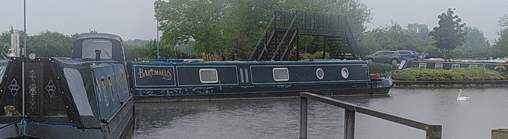 A side view of a narrowboat moored in a marina.  Beyond is a metal bridge to another part of the marina.  Moored boats can be see behind.  