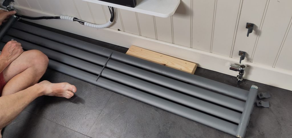 Removed radiator.  The side wall of a narrowboat with brackets for mounting a radiator.  The radiator is lying on the floor beneath.  A man is sitting near the radiator, his crossed bare legs are visible.