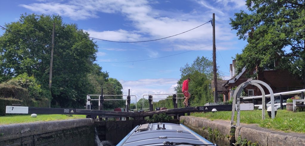 Lock worker. A man in red shorts and jumper is crossing the head gates of a lock.  The roof of the boat rising in the lock is visible in the foreground.  The sky is mainly blue with light fluffy clouds.
