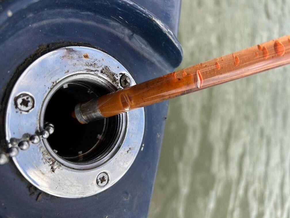 Fuel Bug.  The open fuel filler at the stern of a narrowboat has a plastic tube being removed from it.  The tube contains sampled fuel which is clouded with fluffy looking clumps.