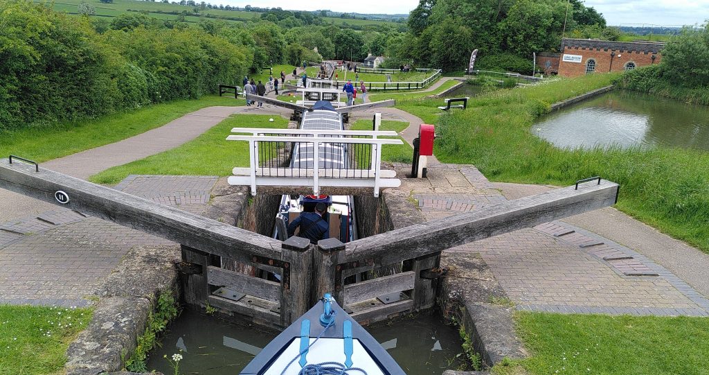 Foxton locks. The bow of a narrowboat is resting on the gates of the lock staircase at Foxton.  The lock below has a narrow bridge across it.  Under the bridge, the roof of another narrowboat can be seen.