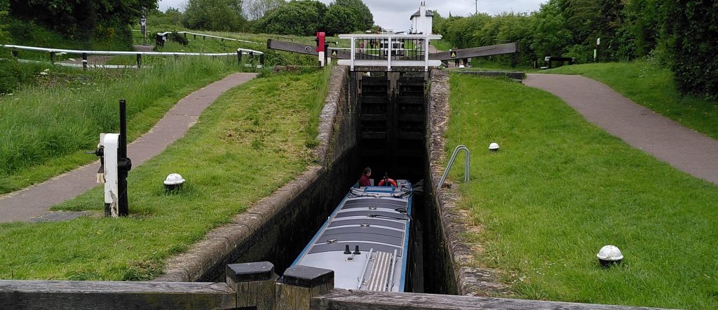 Narrowboat in a staircase.  The narroboat is at the bottom of a lock in a staircase.  The entire boat is below ground level.  The stern is in the sloping section below the massive gates to the lock above and behind.
