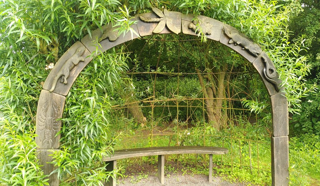 Bower.  A small seat in the woods has a wooden arch over and in front of it.  The arch has large carvings of various things you would find in or near a canal: otter, leaf, pike, eel etc.