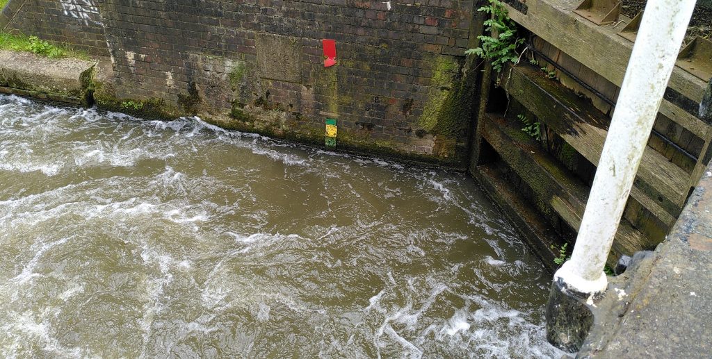 Level marker.  Water is swirling out of a lock gate.  On the stone wall at the far side of the canal is a level gauge.  Just above water level is a short section of green, below which is a short yellow piece.  Marks in the stone show where a red marker previously ran, but only a short part at the top is still in place.